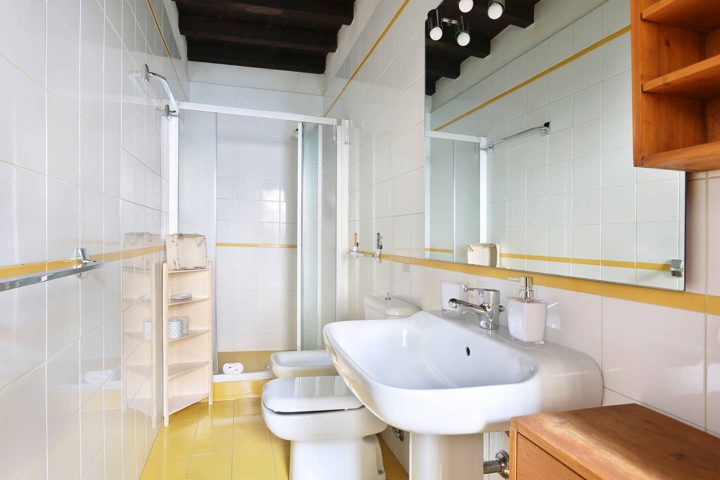 Een badkamer met witte tegels, een gele vloer, een wastafel, toilet, douche en een houten plank.