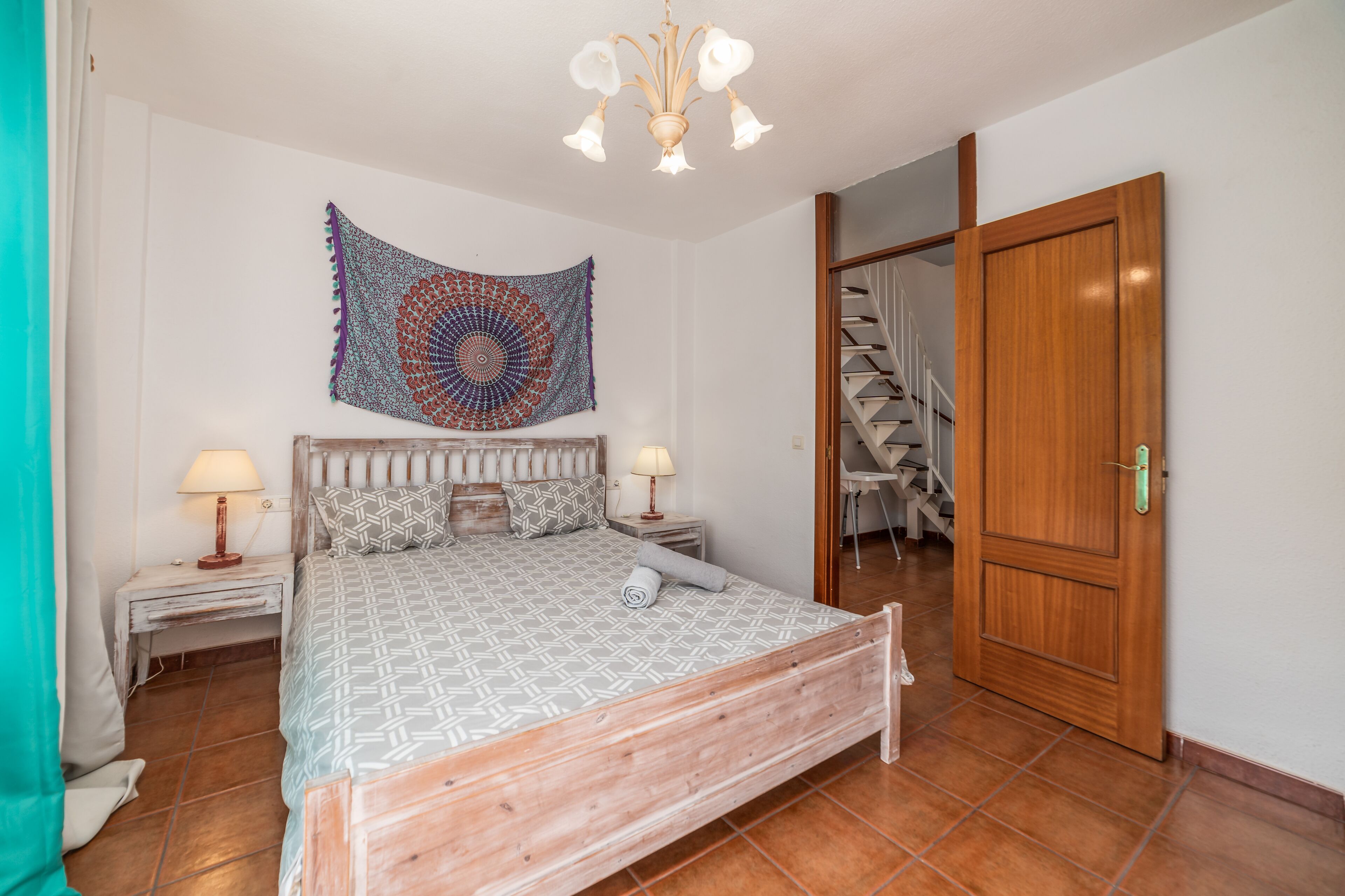 A bedroom with a wooden bed, a patterned bedspread, a tapestry on the wall, a staircase, and a wooden door.