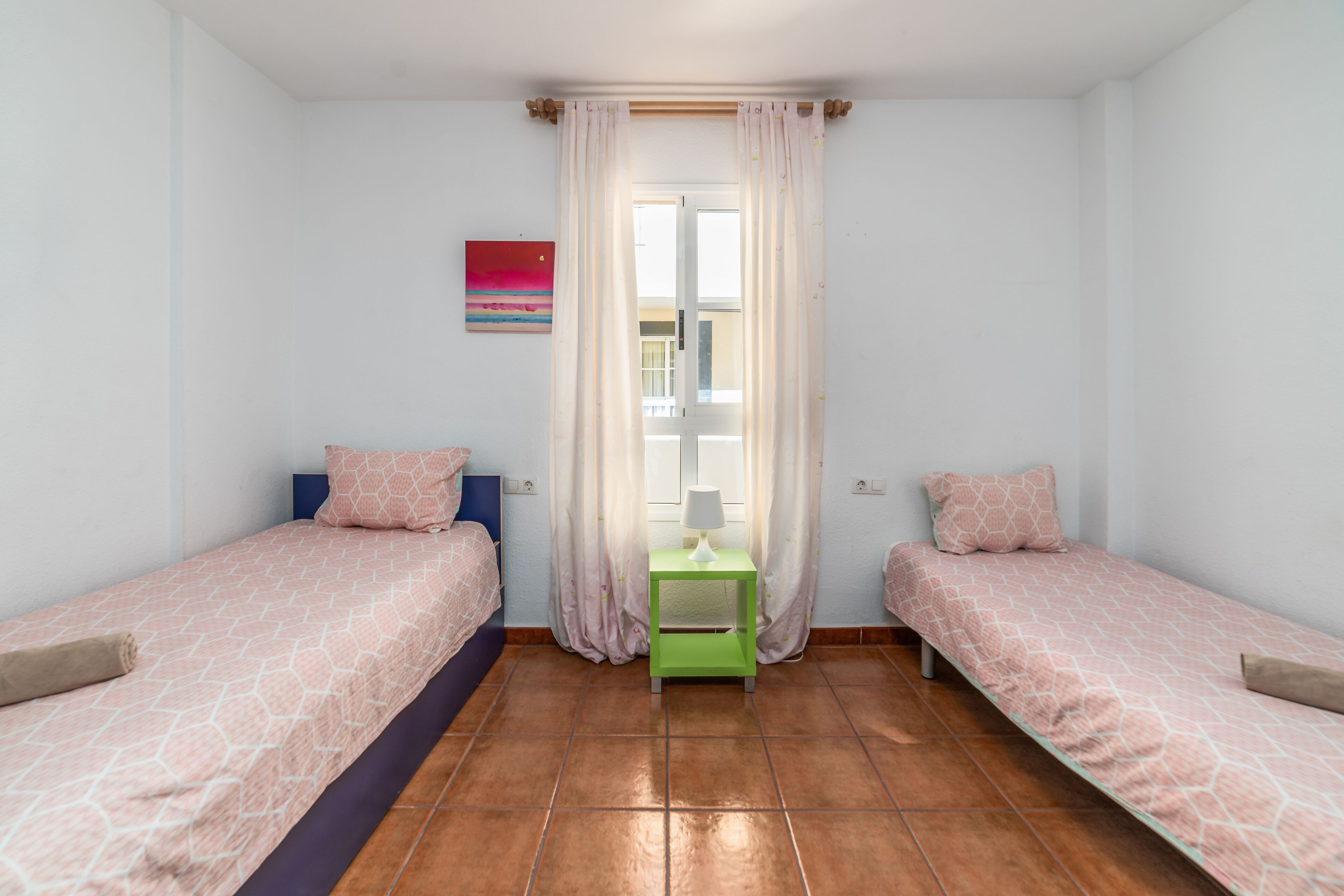 A small, clean room with two beds, a green nightstand, and a window with curtains.
