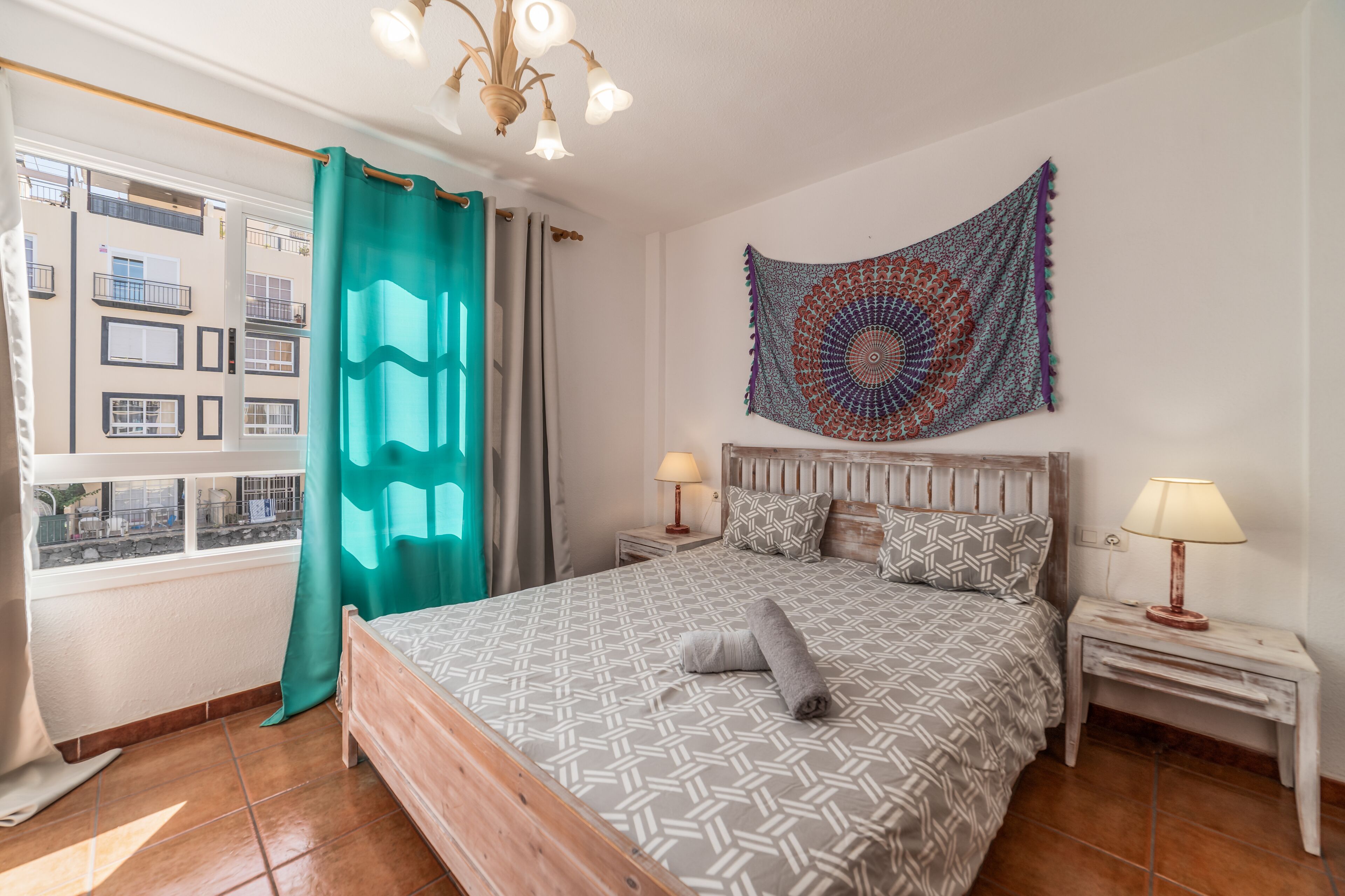 A bedroom with a patterned bedspread, a headboard, two bedside tables with lamps, a tapestry on the wall, and a window with curtains.