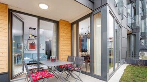 Un área de comedor exterior moderna con una mesa y sillas, ventanas amplias y vistas del interior.