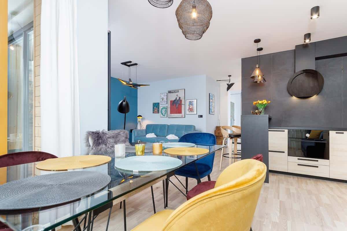 A modern living room with a glass dining table, blue and yellow chairs, a blue sofa, and a kitchen area with a dark backsplash and white cabinets.