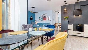 Una sala moderna con una mesa de comedor de vidrio, sillas azules y amarillas, un sofá azul y una zona de cocina con salpicadero oscuro y armarios blancos.