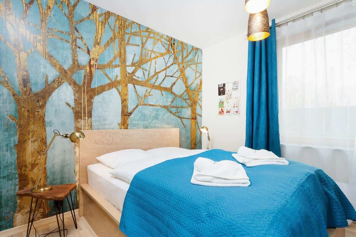 Habitación de hotel con una cama grande, una colcha azul acolchada, un cabecero de madera, un mural de un árbol en la pared y una ventana con cortinas azules.