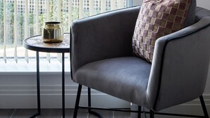 A modern room with a grey armchair, a small round table with a decorative item, and a large window with vertical blinds.