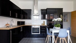 A modern kitchen with black cabinets, a stainless steel range hood, and a dining area with a white table and blue chairs.