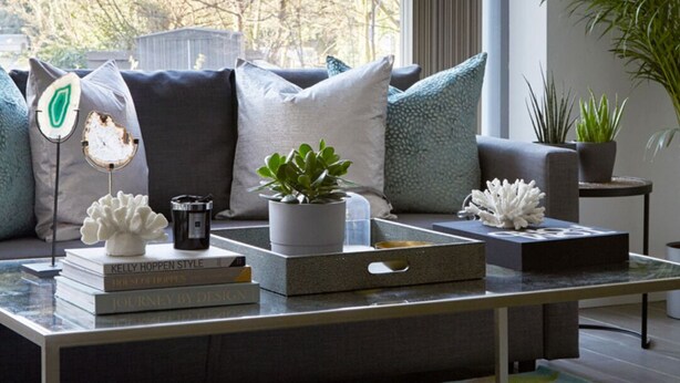 A modern living room with a large window, a sofa with cushions, a coffee table with books and a plant, and a decorative light fixture.