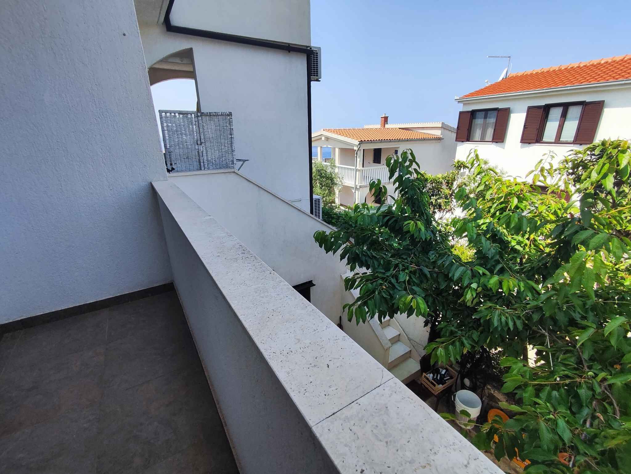 Ein Balkon mit Betonbrüstung, Metallgeländer und Blick auf angrenzende Gebäude und Grünflächen.