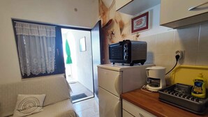 Una cocina compacta con refrigerador, cafetera y tostadora. Hay un sofá con un cojín decorativo y una ventana con cortinas translúcidas.