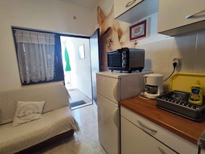 A compact kitchen with a refrigerator, a coffee maker, and a toaster. There is a sofa with a decorative pillow and a window with sheer curtains.