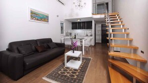 A modern living room with a black leather sofa, a white coffee table, and a patterned rug. A staircase with wooden steps leads to an upper level.