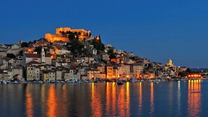 A coastal cityscape at dusk with a prominent hilltop fortress, a harbor with boats, and a domed building.