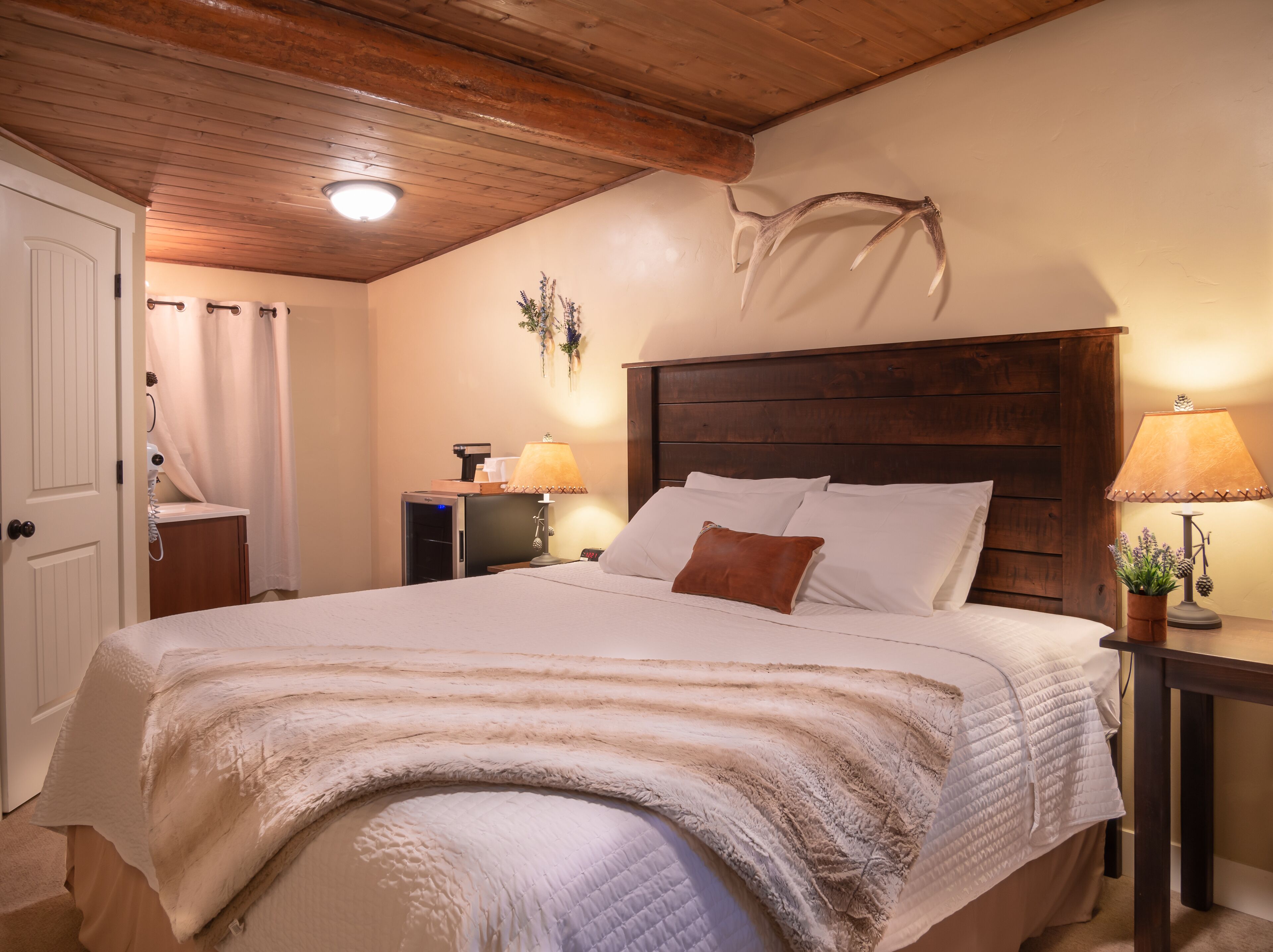 A bedroom with a wooden headboard, a bed with white linens, a nightstand with a lamp, and a wall-mounted deer antler decoration.