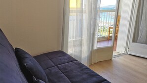 A room with a dark-colored sofa, a sliding glass door leading to a balcony, and a wooden floor.