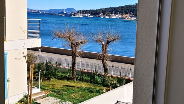 A view from a balcony overlooking a marina with boats, a clear blue sky, and a hilly landscape in the distance.