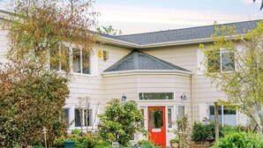A two-story building with a red door, surrounded by a garden with various plants and flowers.