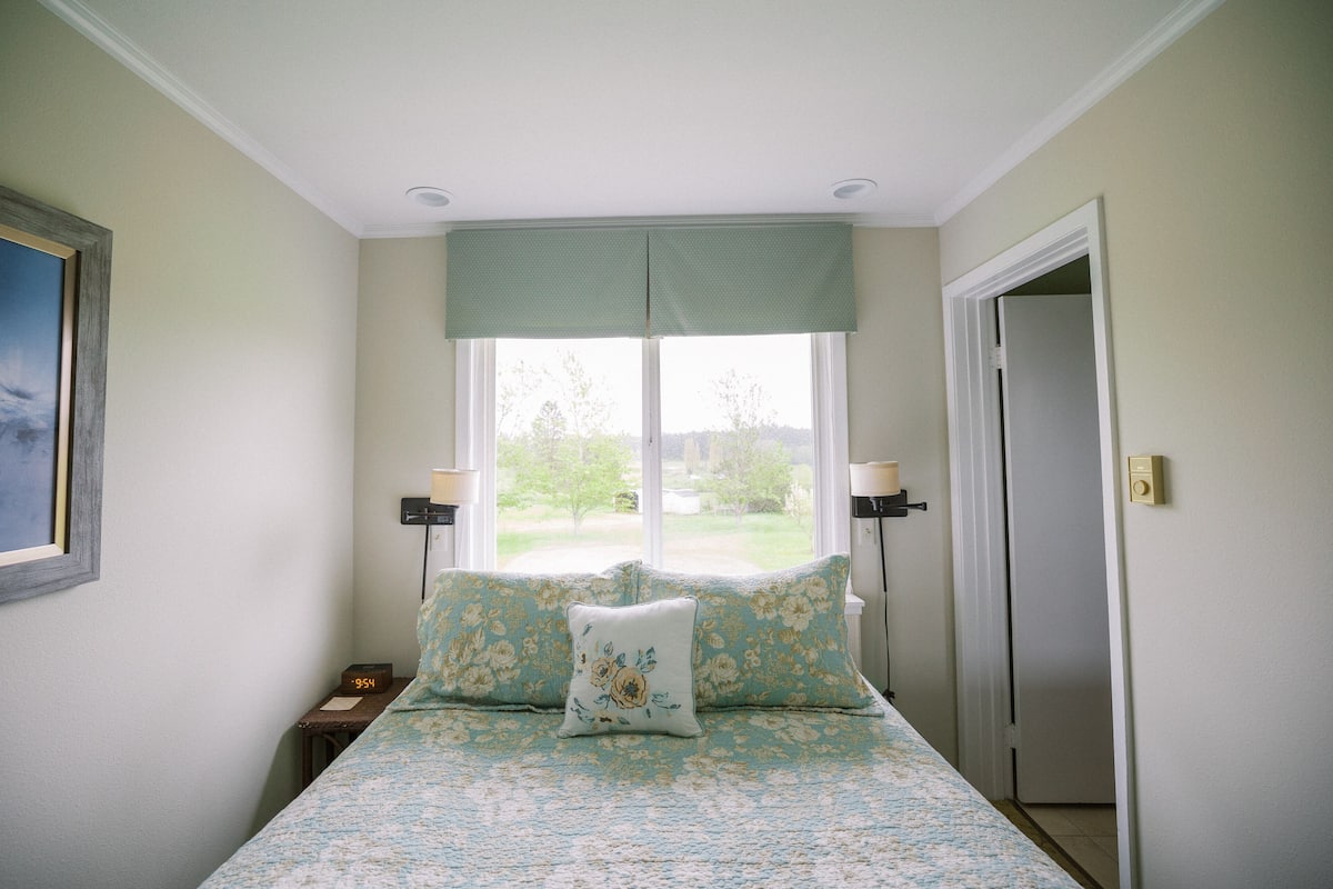 A bedroom with a bed, two bedside lamps, a window with a view of greenery, and a door.