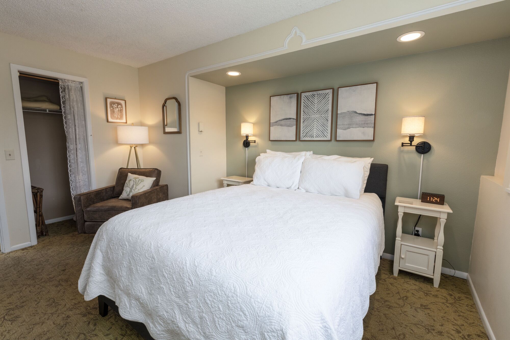 A neatly made bed in a hotel room with a closet, two bedside lamps, and framed artwork on the wall.