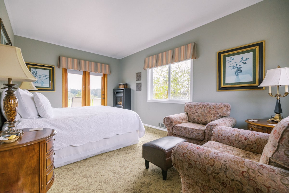 A bedroom with a large bed, two armchairs, a wooden nightstand, a lamp, and a window with curtains.