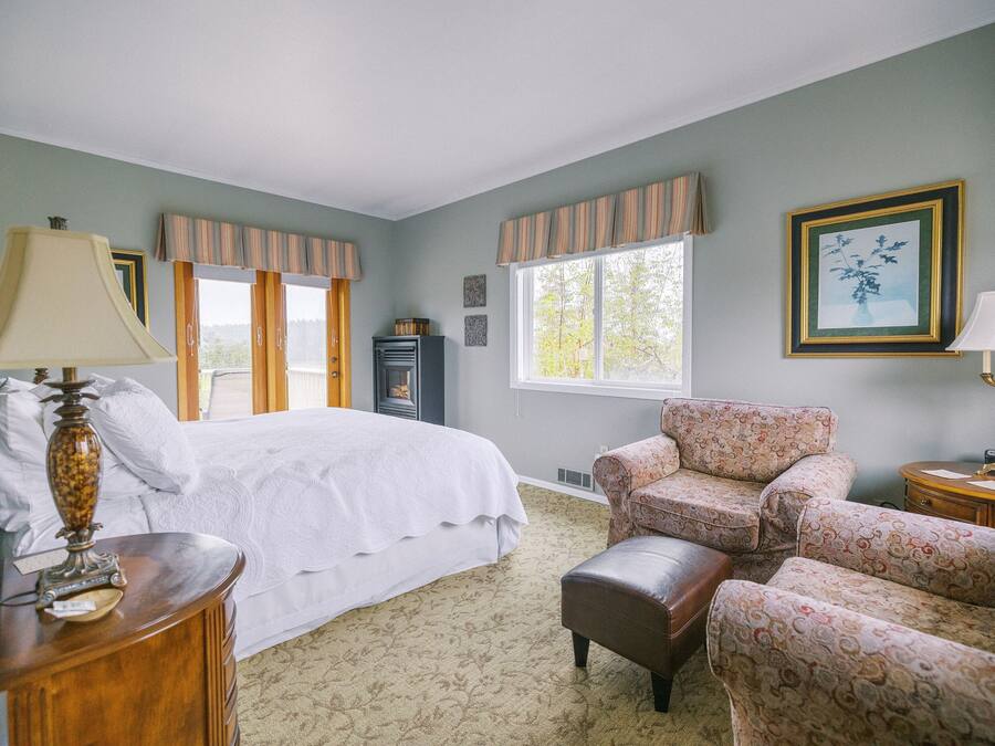 Un dormitorio con cama, dos sillones, una mesita de noche, lámpara, cómoda y ventana con cortinas.