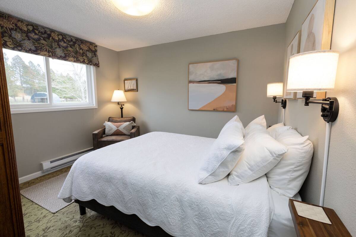 A neatly arranged bedroom with a bed, bedside lamps, a chair, and a window with a view.