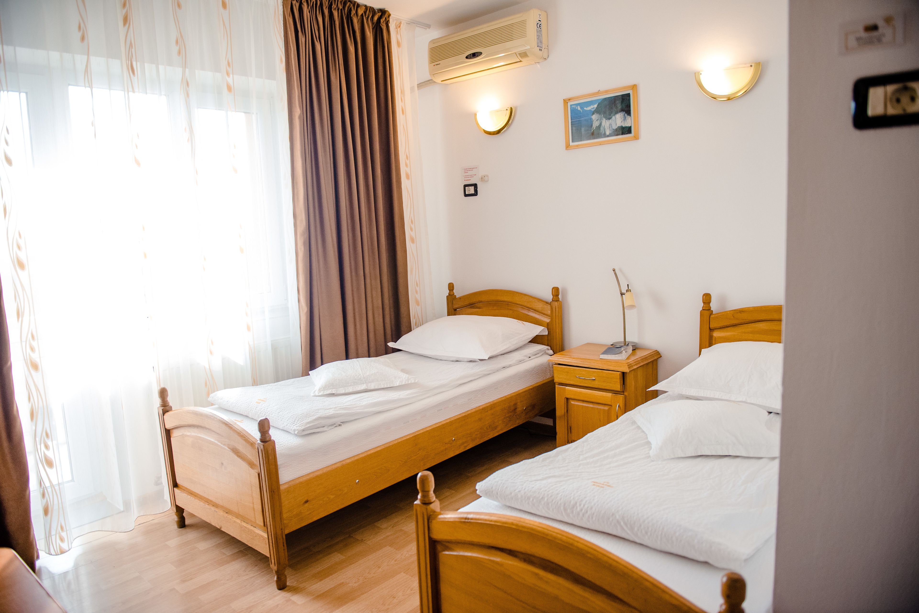 Habitación de hotel con dos camas de madera, una mesita de noche con lámpara, aire acondicionado y ventana con cortinas.