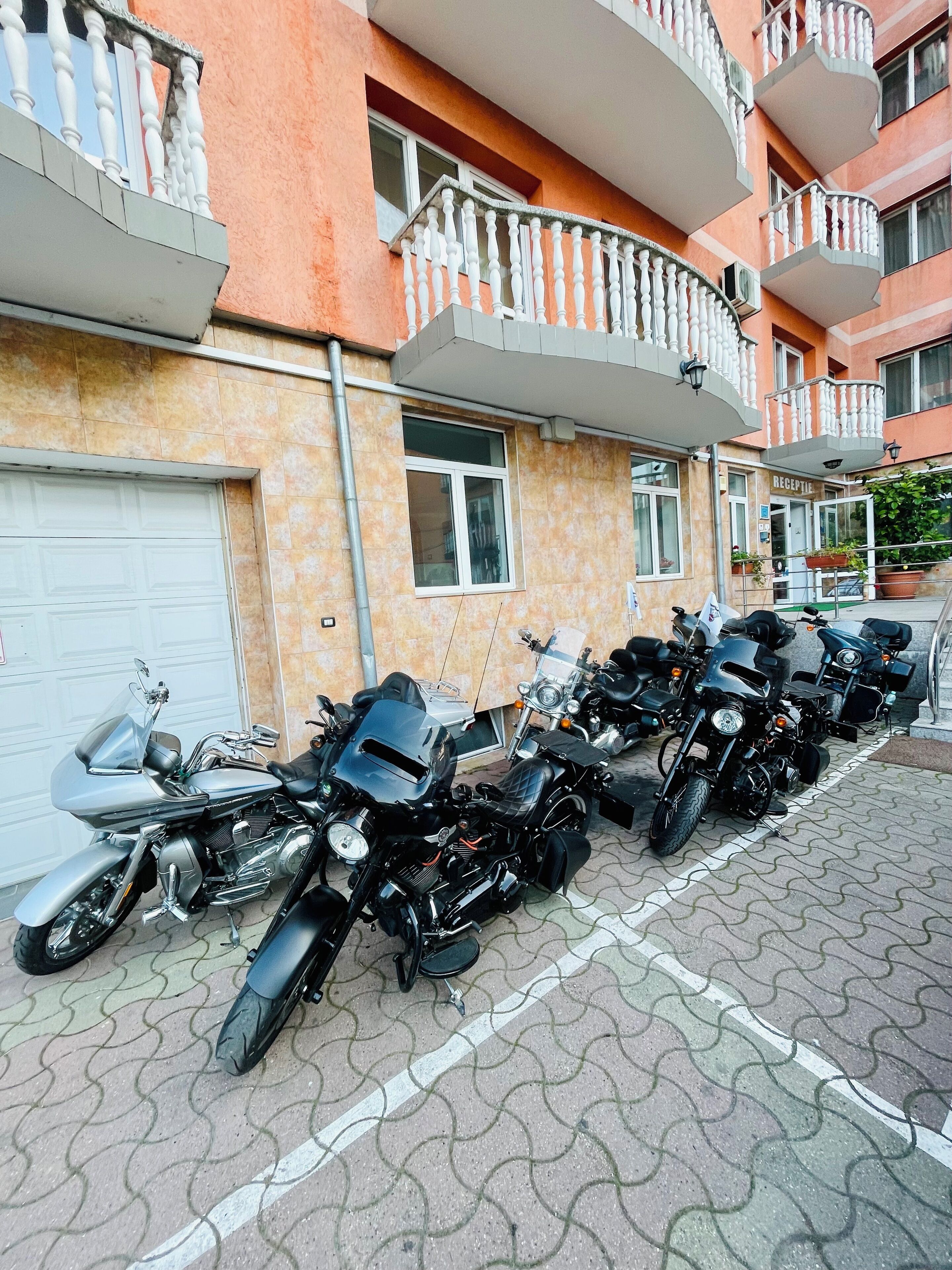 Rivi parkkeerattuja moottoripyöriä rakennuksen edessä, jossa on laatoitettu piha-alue ja valkoisilla kaiteilla varustettu parveke.