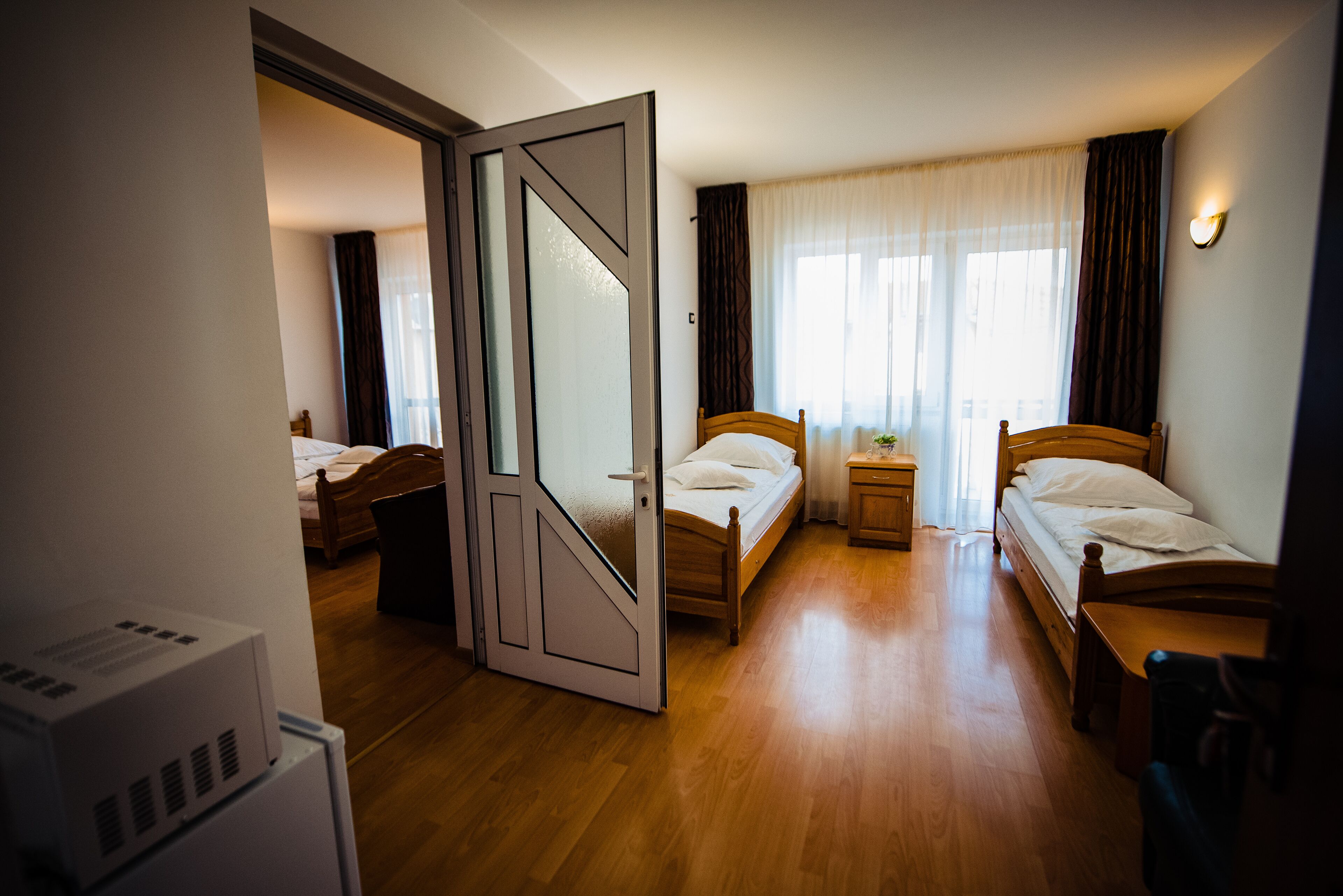 Une chambre d’hôtel avec deux lits simples, un plancher en bois, une porte ornée de motifs géométriques et une table de chevet avec une lampe.