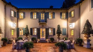 Une cour avec des espaces de repos en plein air, des plantes en pot et un bâtiment doté de nombreuses fenêtres et volets.