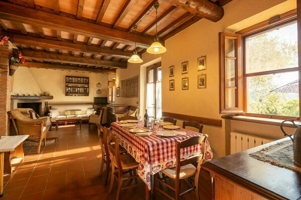 Un coin repas rustique avec une table en bois dressée pour quatre personnes, entourée de chaises en osier. On aperçoit une cheminée au fond de la pièce, et le plafond est orné de poutres apparentes.