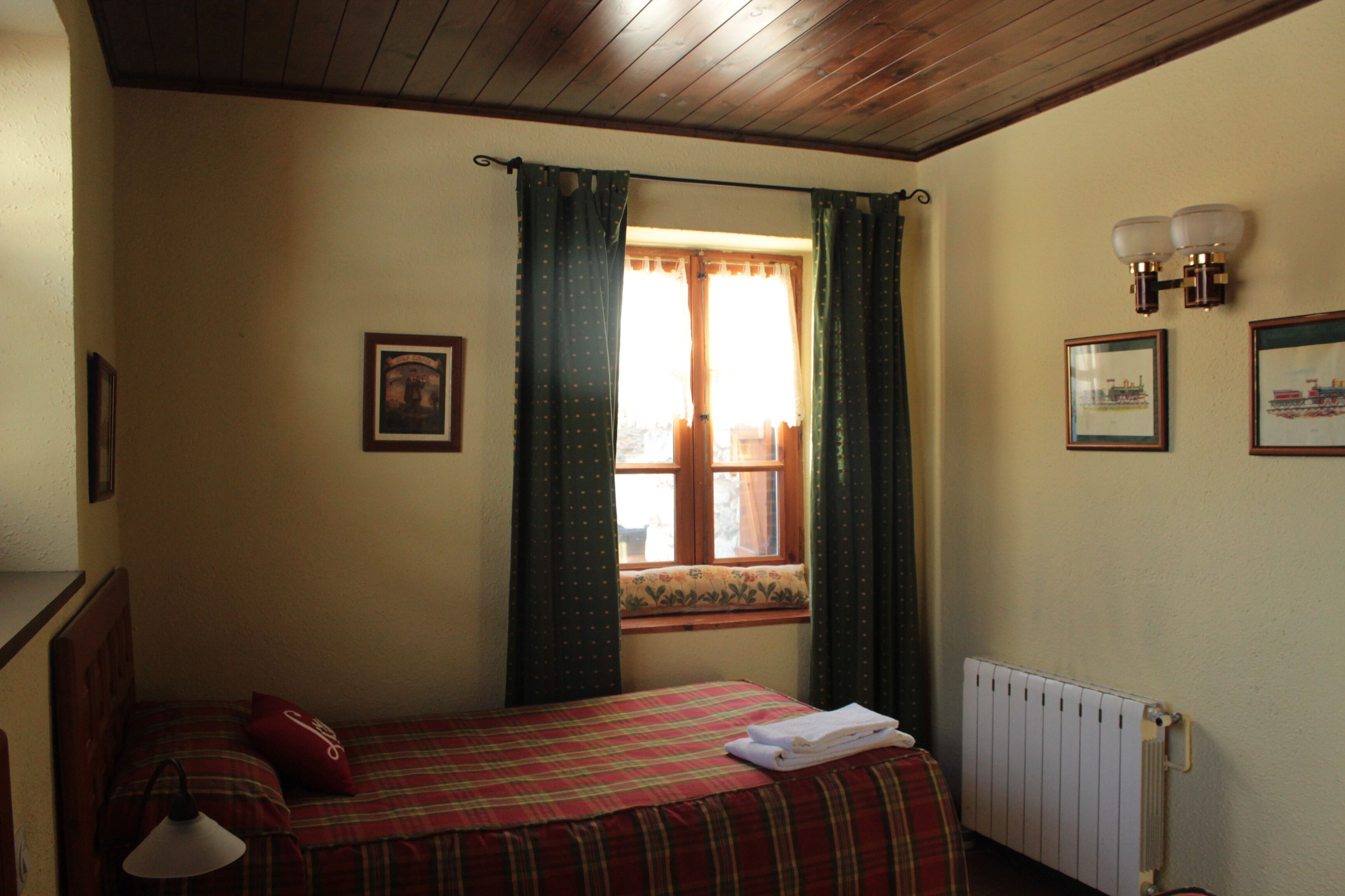 Un dormitori amb llit, finestra amb cortines, radiador i llums penjades a la paret.