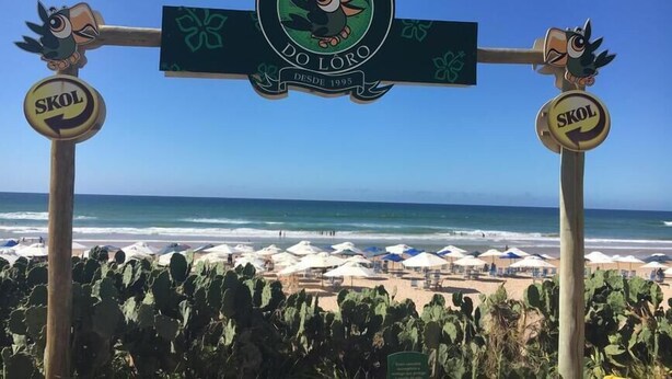 Blick auf einen Strand, mit einem Schild für 'Barraca do Loro' und Schildern für 'SKOL'.