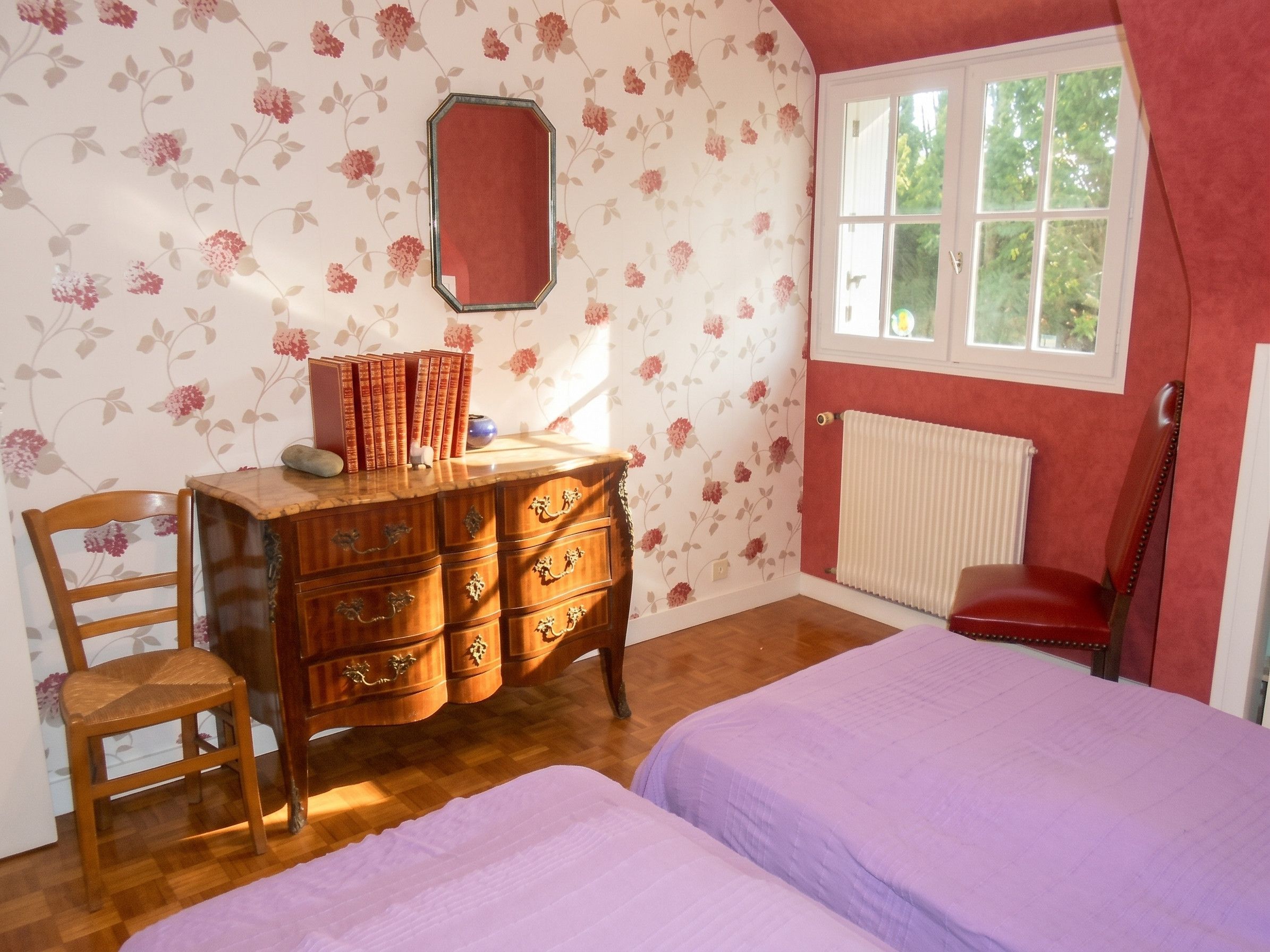 A bedroom with a wooden dresser, two beds, a chair, and a window with a view of greenery.