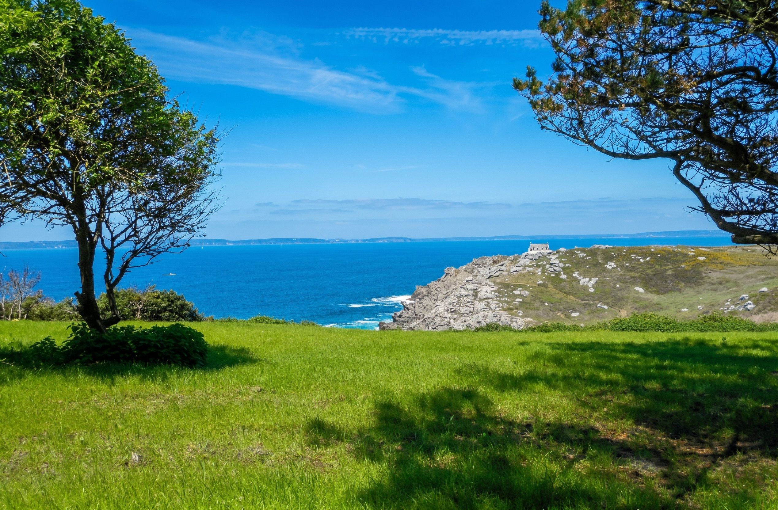 A coastal landscape with a clear view of the sea, a rocky outcrop, and a small building on the right.