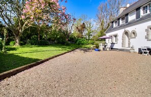 A gravel driveway leading to a white building with stone accents, surrounded by a well-maintained garden with flowering trees.