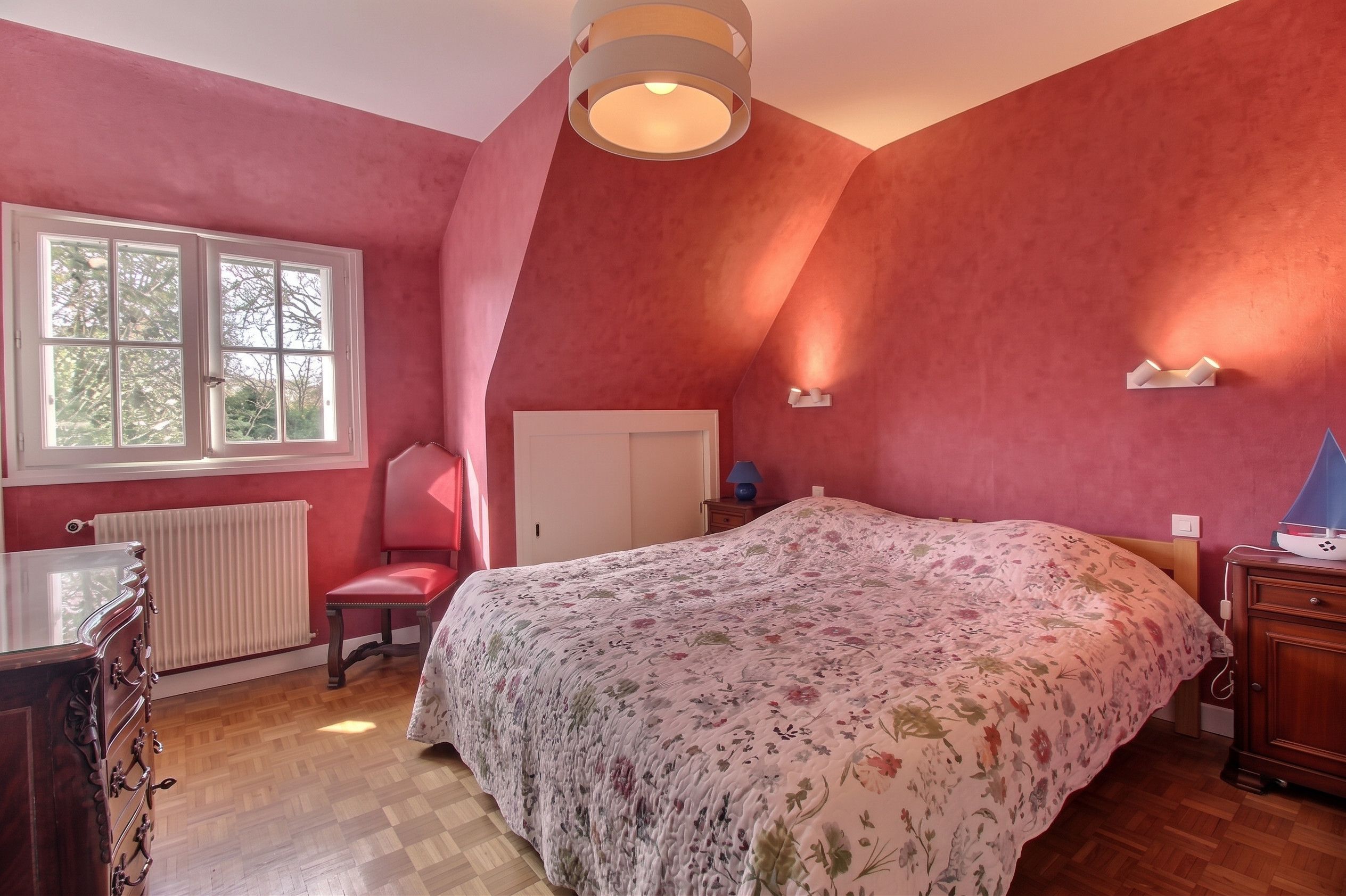 Ein Schlafzimmer mit einem großen Bett, einem Sessel, einem Schreibtisch, einer Heizung und einem Fenster mit Blick auf Bäume.