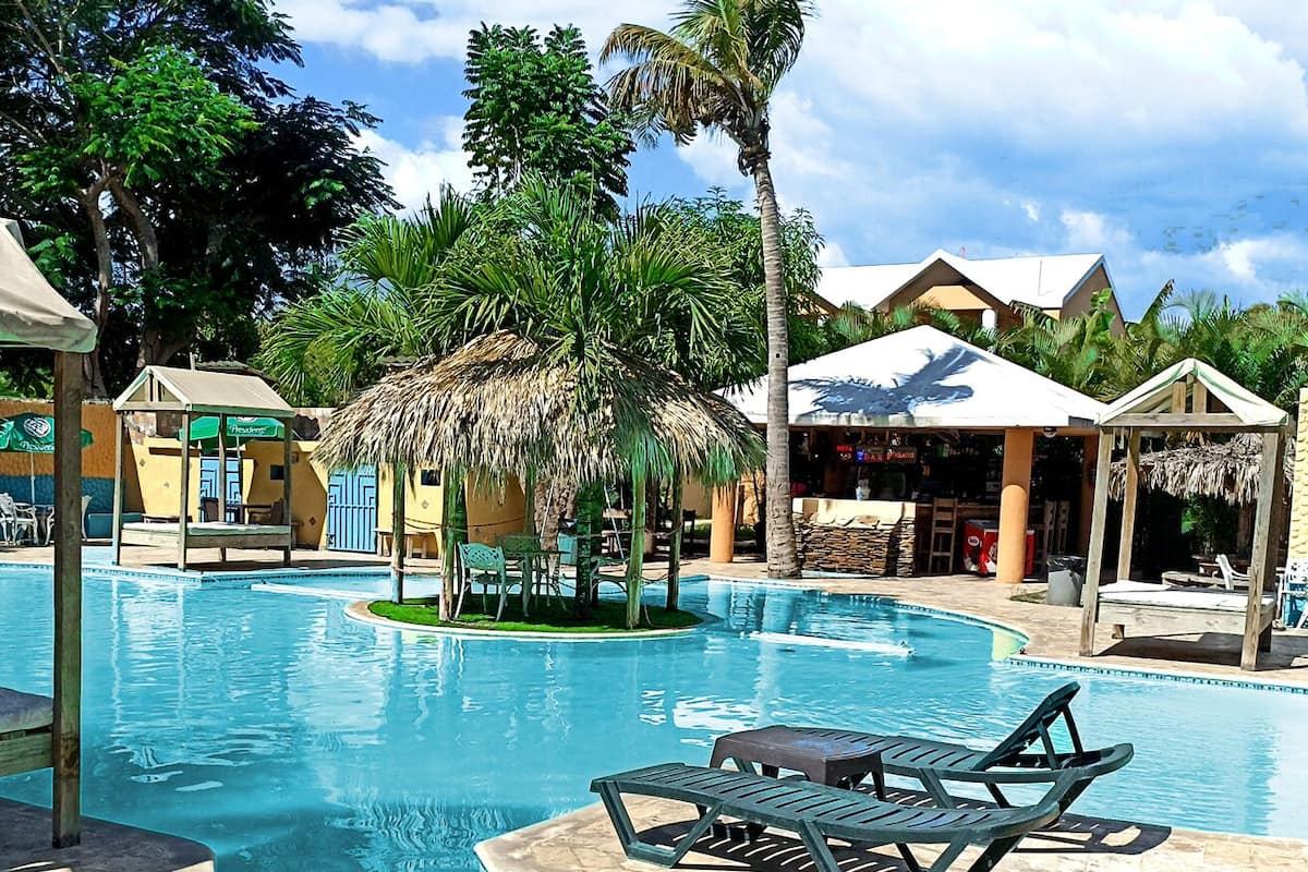 A resort pool area with lounge chairs, a thatched-roof structure, and palm trees.