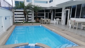 Una piscina con solárium de baldosas, rodeada de sillas y mesas blancas, y palmeras.