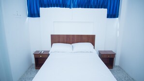 Una cama bien tendida con ropa blanca, dos mesitas de noche de madera, con un control remoto sobre una de ellas, y una cortina azul sobre la ventana.