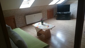 Ein Zimmer mit Schlafgalerie, Holzboden, einem grünen Sofa, einem kleinen runden Tisch mit Blumenmuster und einem Heizkörper. Ein Fernsehschrank ist ebenfalls vorhanden.