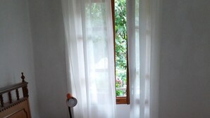 Una cama bien hecha con toallas dobladas, cabecera de madera, lámpara de noche y ventana con cortinas traslúcidas.