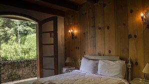 Dormitorio con paneles de madera, una cama, una mesita de noche, una lámpara y vistas al exterior con vegetación.
