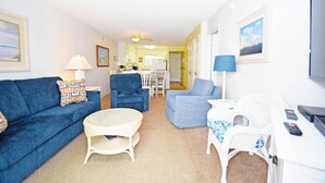 Un salon avec un canapé bleu, un fauteuil blanc et une chaise en osier. Une petite table ronde se trouve au centre de la pièce.