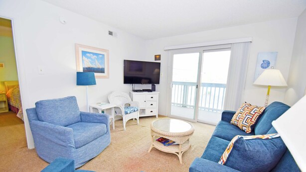 Un salon avec un canapé bleu, un fauteuil blanc, une table basse ronde, un téléviseur à écran plat et un balcon doté d’une balustrade blanche.