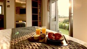 Una cama bien hecha con colcha, una bandeja con cruasanes y jugo, y una estantería al fondo.