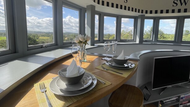 Un comedor con una mesa lista para dos personas, ventanas amplias y un televisor.