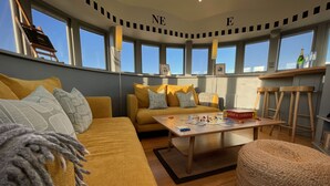 Un salón acogedor con un sofá amarillo, una mesa de centro con juegos de mesa y ventanas amplias con vistas.