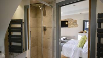 A modern bathroom with a glass shower enclosure, a towel rack, and a visible bed with a green pillow in the adjacent room.