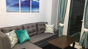 Un salon avec un canapé gris, deux coussins à motifs, une petite table en bois et une porte-fenêtre coulissante avec des rideaux.