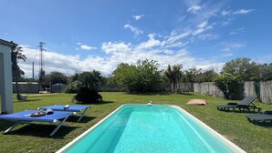 Una piscina rectangular con agua azul cristalina, rodeada por un césped con valla de madera, árboles y mobiliario exterior, incluyendo sillones de descanso y una mesa.